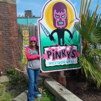 A woman (Nancy) stands beside the outdoor sign for Pinky's Westside Grill in Charlotte, NC, smiling at the camera. The illustrated sign features a cartoon luchador wrestler character holding a knife against a yellow and green circular background with the restaurant name in bold lettering. She is wearing a pink baseball cap, sunglasses, a Star Wars hoodie, and jeans. A brick wall, colorful murals, and palm-like plants are visible in the background.