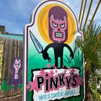 The outdoor sign for Pinky's Westside Grill in Charlotte, NC. The large illustrated sign features a cartoon luchador wrestler character wearing a dark purple mask and holding a knife, set against a bright yellow and green circular background. The restaurant name "Pinky's Westside Grill" is displayed in bold lettering at the bottom. Colorful murals and desert plants are partially visible on either side of the sign.