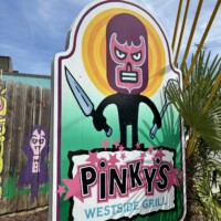 The outdoor sign for Pinky's Westside Grill in Charlotte, NC. The large illustrated sign features a cartoon luchador wrestler character wearing a dark purple mask and holding a knife, set against a bright yellow and green circular background. The restaurant name "Pinky's Westside Grill" is displayed in bold lettering at the bottom. Colorful murals and desert plants are partially visible on either side of the sign.