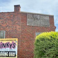 The exterior brick wall of Pinky's Westside Grill in Charlotte, NC, showing a faded ghost sign painted directly on the upper portion of the building, barely legible against the red brick. A bold yellow and black "This Field is Reserved for Pinky's Westside Grill Parking Only" sign is mounted on the lower portion of the wall. A trimmed yellow-green shrub is visible at the lower right corner.