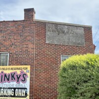 The exterior brick wall of Pinky's Westside Grill in Charlotte, NC, showing a faded ghost sign painted directly on the upper portion of the building, barely legible against the red brick. A bold yellow and black "This Field is Reserved for Pinky's Westside Grill Parking Only" sign is mounted on the lower portion of the wall. A trimmed yellow-green shrub is visible at the lower right corner.
