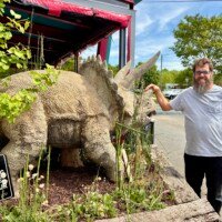A man (Zach) stands beside a life-sized concrete triceratops dinosaur sculpture in the outdoor area of Pinky's Westside Grill in Charlotte, NC, grinning and pointing at it. A small handwritten sign in front of the planter reads "No Climbing on the Dinosaur." The dinosaur is surrounded by green plants and wildflowers in a raised garden bed. The restaurant's exterior with a red awning is partially visible in the background.