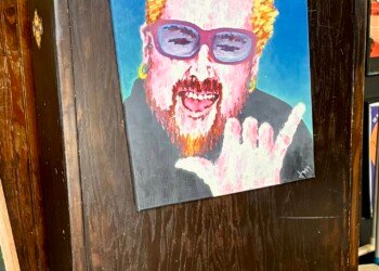 A painted portrait of Guy Fieri mounted on the front of a dark wooden host stand inside Pinky's Westside Grill in Charlotte, NC. The colorful illustration depicts him with his signature spiky bleached hair, blue-tinted sunglasses, and red beard, flashing a rock and roll hand gesture and grinning widely. The walls behind are covered in framed artwork, posters, and stickers typical of the restaurant's eclectic interior decor.