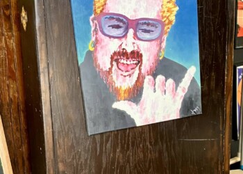 A painted portrait of Guy Fieri mounted on the front of a dark wooden host stand inside Pinky's Westside Grill in Charlotte, NC. The colorful illustration depicts him with his signature spiky bleached hair, blue-tinted sunglasses, and red beard, flashing a rock and roll hand gesture and grinning widely. The walls behind are covered in framed artwork, posters, and stickers typical of the restaurant's eclectic interior decor.