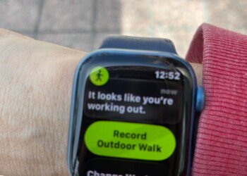 A close-up of an Apple Watch on a woman's wrist displaying a workout detection notification. The screen reads "It looks like you're working out." with a bright green button below it saying "Record Outdoor Walk" and a secondary option for "Change Workout." The time shows 12:52. A pink sleeve is visible at the wrist and a brick sidewalk is partially visible at the bottom of the frame.