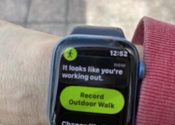 A close-up of an Apple Watch on a woman's wrist displaying a workout detection notification. The screen reads "It looks like you're working out." with a bright green button below it saying "Record Outdoor Walk" and a secondary option for "Change Workout." The time shows 12:52. A pink sleeve is visible at the wrist and a brick sidewalk is partially visible at the bottom of the frame.
