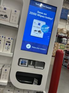 A Freeosk sample kiosk inside a Sam's Club warehouse store, displaying a promotion for ZEISS Lens Wipes on its digital screen. The screen advertises a $1 off coupon and reads "Enjoy your sample!" A sample packet is visible dispensed in the lower tray. Shelves stocked with ZEISS lens care products are visible to the left, and a red warehouse column is partially visible on the right.