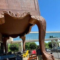 A woman stands near the base of the giant highboy dresser's leg, with patio chairs and a glass walkway visible in the background at Furnitureland South
