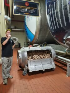 Tour guide demonstrating the beechwood aging process next to an open drum of beechwood chips at Anheuser-Busch, with a sign reading Beechwood Aging Cellars Capacity 3600 Barrels.