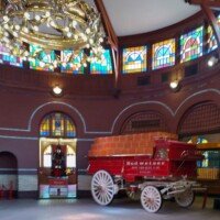 A historic red Budweiser delivery wagon loaded with crates, parked beneath a brass chandelier and stained glass dome inside the Anheuser-Busch Brew House.
