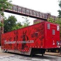 Large red Budweiser Clydesdales horse transport trailer parked beneath an elevated pipe walkway at the Anheuser-Busch brewery.
