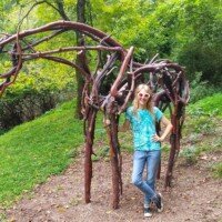 Nancy standing under a large arched sculpture made of interwoven tree branches at Crystal Bridges Museum.