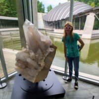 Nancy standing beside a massive quartz crystal cluster on display at Crystal Bridges Museum, with the museum's pond and bridge architecture visible through the window.