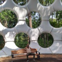 Zach sitting on a wooden bench inside Buckminster Fuller's Fly's Eye Dome, surrounded by trees visible through the circular openings.
