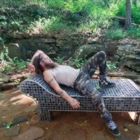 Zach lounging on a stone and wire-mesh chaise bench along a Crystal Bridges trail, surrounded by moss-covered rock walls.