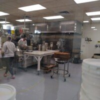 View through a glass window into the candy kitchen, with workers in hairnets, a marble-top table, a copper kettle and colorful scoops hanging overhead