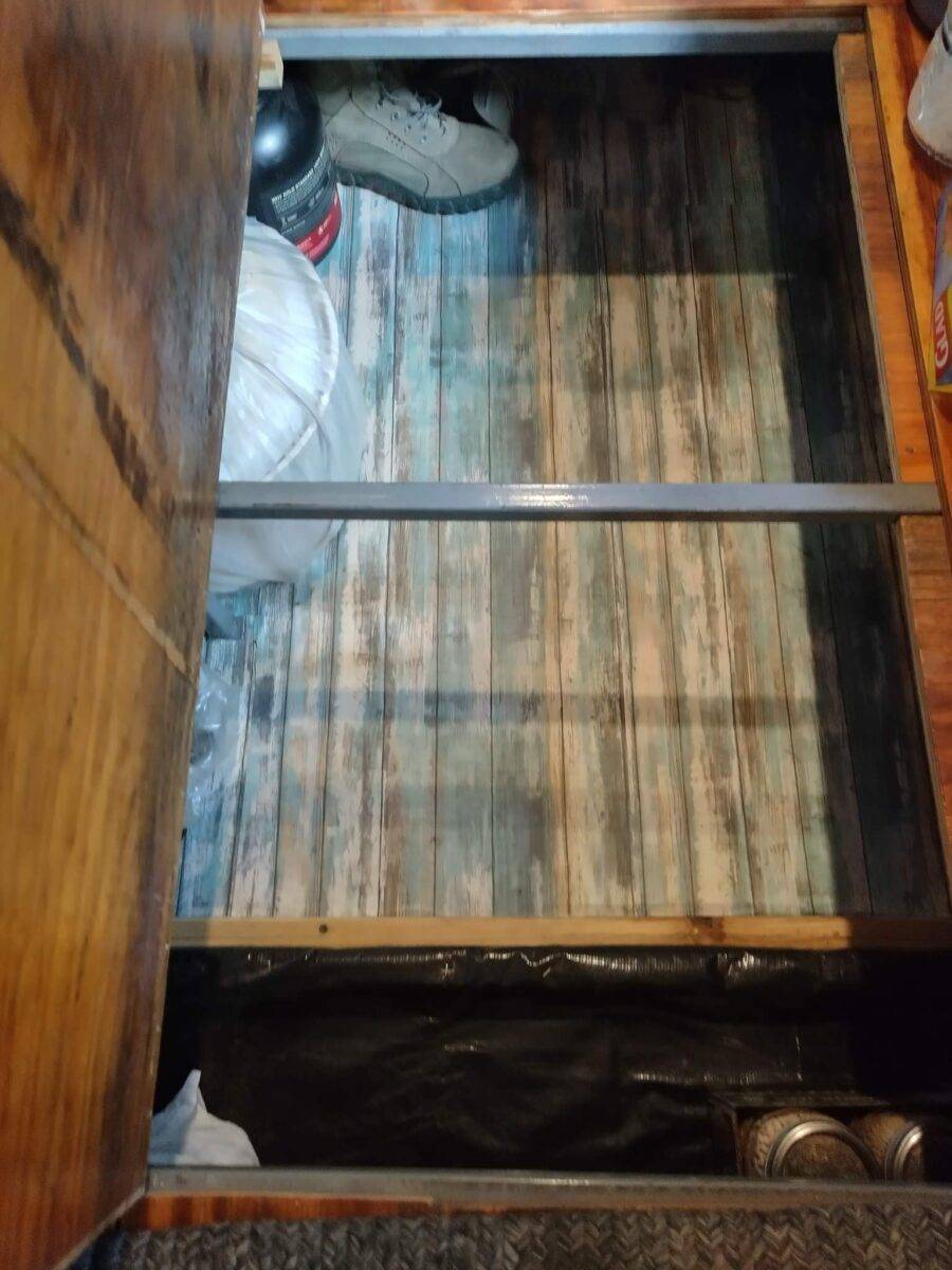 Under-floor storage compartment in converted bus revealed by lifted wood panel, containing shoes, helmet, and black cylindrical object, with striped surface inside