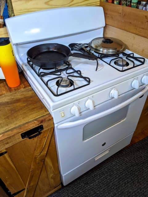 Full-size white gas stove in converted bus kitchen with frying pan, covered pot, wooden cabinets, and countertop with yellow bottle, all set against a carpeted floor.