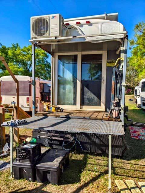 Open back deck attached to converted bus with metal railing, small table, storage bins, and sliding glass doors leading into the interior.