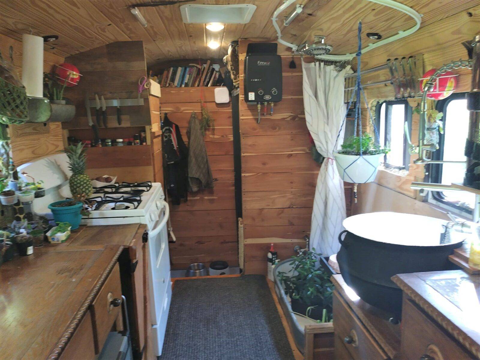 Interior of converted bus kitchen with full-size white stove, wood-paneled walls and ceiling, countertop with produce, bookshelves above, and potted plants beside sink and window