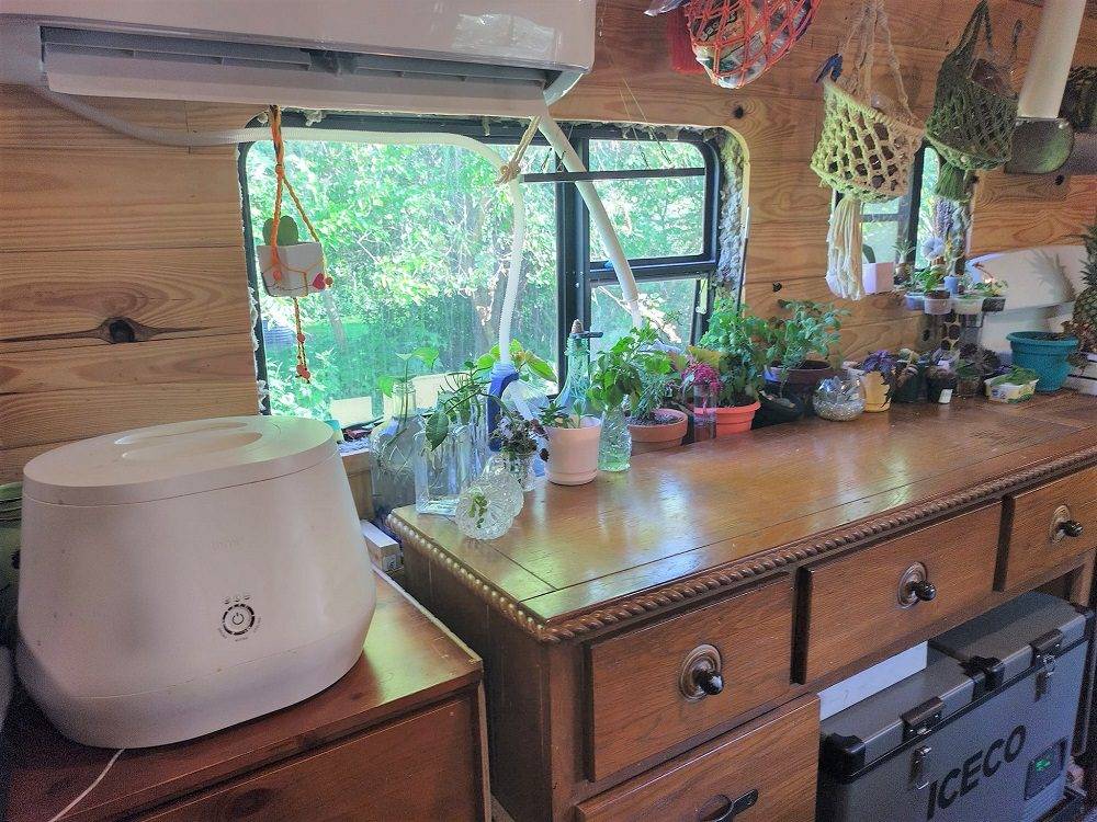Converted bus interior with wooden dresser topped with potted plants and decor, ICECO refrigerator below, air conditioning unit above curtained window, and hanging net organizer on right wall.