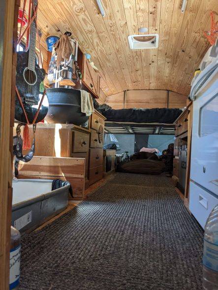 Converted bus kitchen with wood-paneled ceiling, carpeted floor, hanging black pot, wooden cabinetry on both sides, and compact white appliance beside drawers