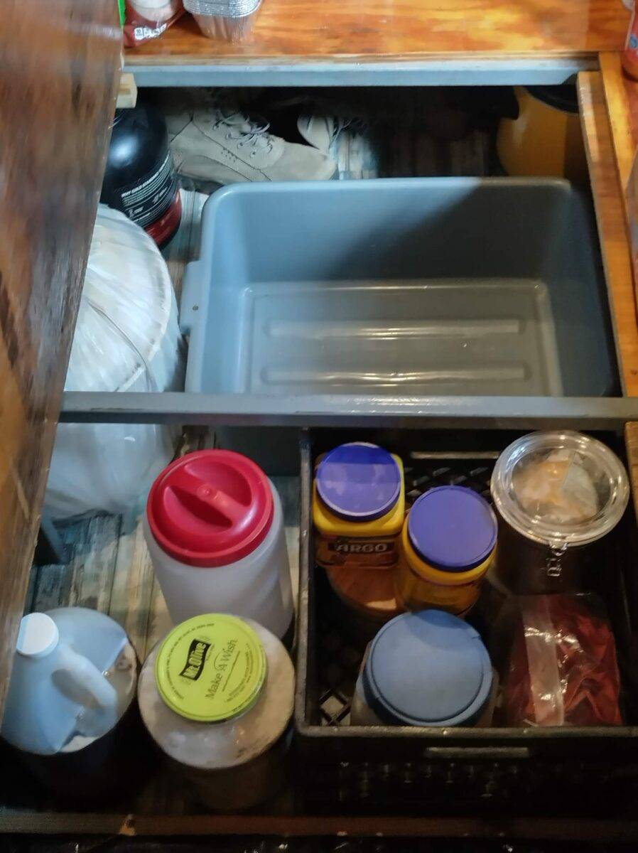 Organized under-compartment bus storage with various plastic containers, mason jar, white jug, packaged food, and cleaning supplies inside black bin and gray tub