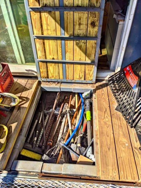 Open deck storage compartment on converted bus containing hand tools including broom, hammer, hacksaw, crowbar, pipe wrench, and other tools organized in wooden enclosure