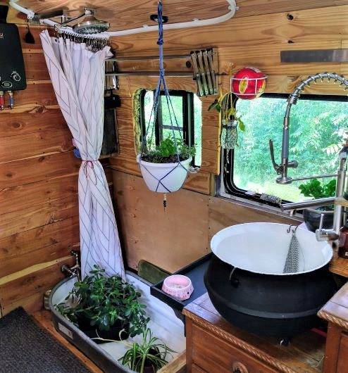Bus kitchen sink area with black basin, silver faucet, wooden cabinetry, hanging plant, pink soap dish, and window with white curtain above small indoor tub containing potted plants.