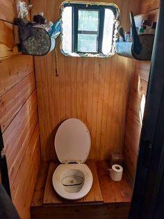 Compact bathroom inside converted bus with wood-paneled walls, open composting toilet, small window, and shelves holding toiletries and a metal container.