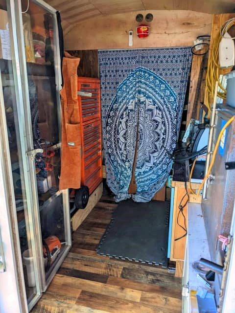 Small organized tool area inside converted bus—orange tool chest, hanging storage, and curtain leading to workshop nook.