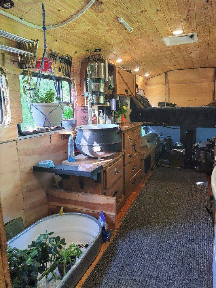 Converted bus kitchen with wooden cabinetry, sink under curtained window, produce basket, countertop appliances, and a view into built-in shelving and sleeping loft above driver’s area.