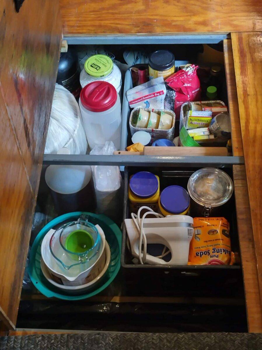 Open floor cabinet in converted bus containing stacked bowls, jars, small kitchen appliances, packaged foods, plastic containers, and rolled paper products