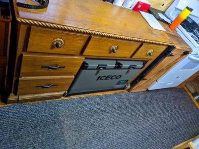Converted bus kitchen with white stove in background, wooden dresser in foreground containing ICECO portable refrigerator behind open compartment, on carpeted floor.
