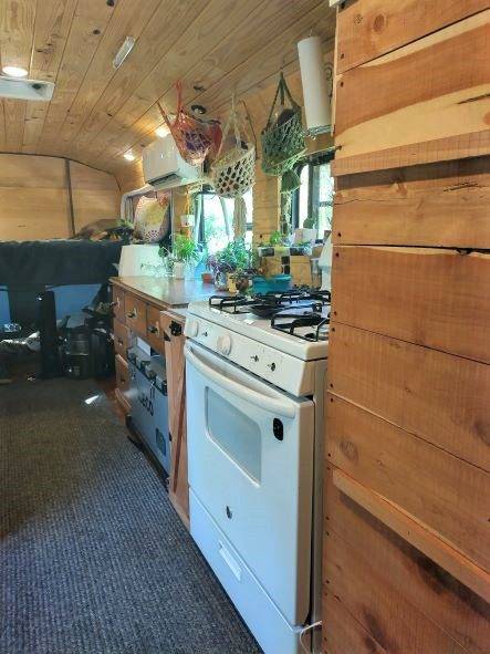 Converted bus kitchen with full-size white stove, wood countertops, overhead cabinets with baskets, hanging plants by curtained windows, and wood-paneled ceiling.