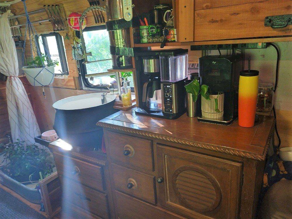 Bus kitchen interior with wooden drawers, countertop appliances including coffee maker, open shelves with jars, and white curtain above small sink under window