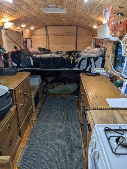 Converted bus kitchen featuring full-size propane range and cast iron cauldron sink set into wooden countertop beneath curtained window.